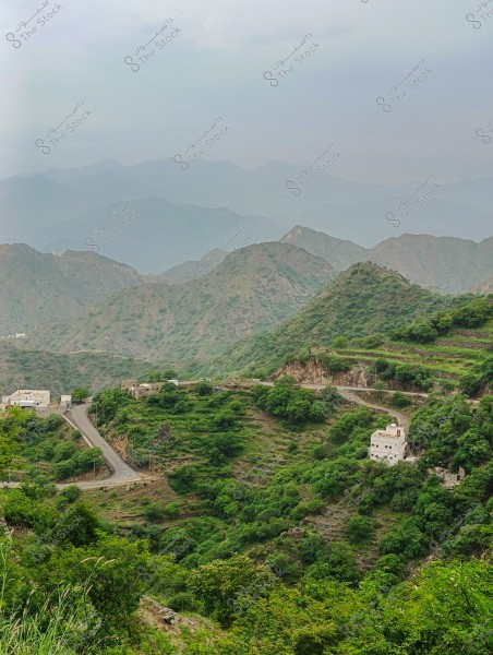 A natural landscape featuring a mountain range with winding roads and small white buildings nestled amidst lush green slopes. The mountains overlap and fade into a pale blue background due to mist and cloudy weather.