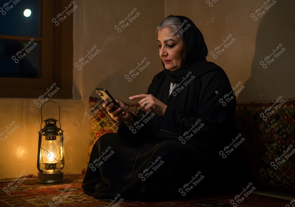 A woman sitting on a traditional carpet beside a lit lantern, dressed in a black abaya and headscarf, looking at a mobile phone in her hand. A window is visible in the background with the moon shining in the sky.