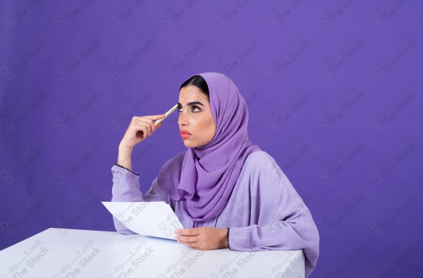 Portrait of a young Saudi woman wearing an abaya and hijab, holding pen and paper and thinking, against a purple background