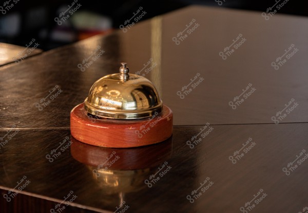 An image of a metallic round service bell on a circular wooden base, placed on a dark glossy desk surface. The bell reflects the surrounding light, emphasizing its sleek and classic shape.