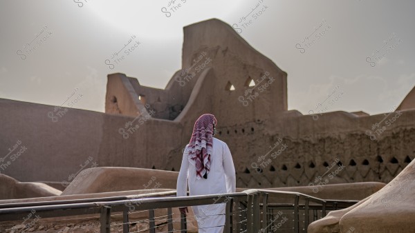 An image of a man wearing a white thobe and a red ghutra walking near a historic building in Saudi Arabia, featuring mud walls and traditional architecture. The sun shines brightly in the sky, casting gentle shadows on the structure.