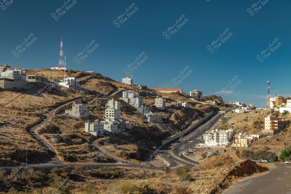 Image of a mountainous city with winding roads featuring multi-story houses spread across barren hills. Several white and beige buildings are clustered on the hills. A tall metal communication tower is visible on one of the peaks. The sky is a clear blue above the mountainous landscape.