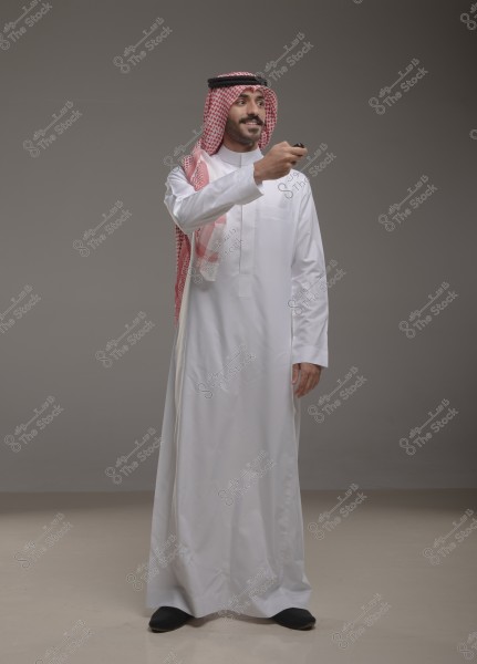 A portrait of a man standing and wearing a traditional white thobe with a red checkered shemagh and agal, likely from Saudi Arabia. The man appears to be smiling and looking sideways, holding a remote control in his hand. The background is a neutral gray color.