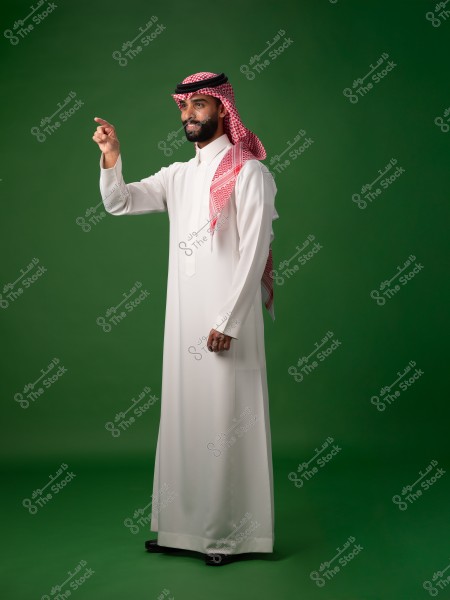 The image shows a man standing against a green background, wearing a white thobe with a red and white checkered shemagh and an agal. He appears to be pointing at something in front of him.