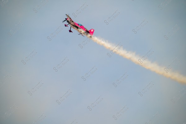 An aerobatic plane performing a maneuver in the blue sky, leaving behind a trail of white smoke resembling a ribbon extending in the direction of flight. The plane is brightly colored in red and white.