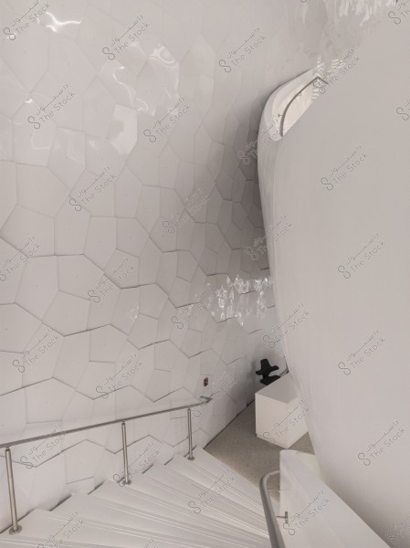 The image shows a modern interior design with a wall covered in white pentagonal tiles. There is a staircase with white steps adjacent to the wall, featuring a metal handrail. In the lower right corner, part of a white desk and a black chair are visible, suggesting a waiting or reception area. The lighting is calm, and the atmosphere is overall modern and minimalist.