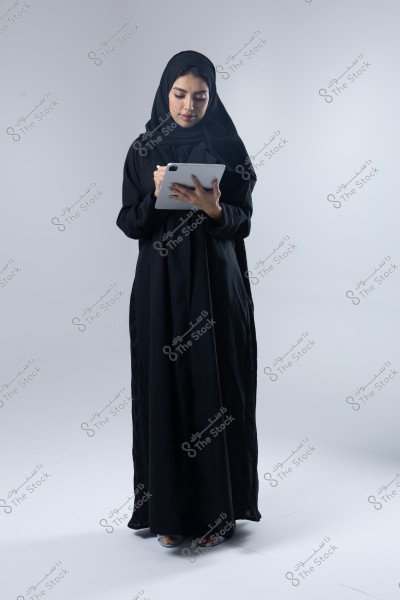 صورة لامرأة ترتدي عباءة سوداء وحجابًا أسود, تقف وتستخدم جهازًا لوحيًا بيدها. يبدو أن الخلفية بيضاء وهادئة, مما يبرز مظهرها التقليدي الأنيق.