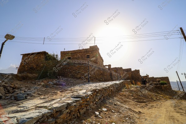 The image depicts old stone buildings situated on a hill, with a bright sun in the background. The setting appears to be a dry, mountainous area. There is a small paved path alongside the buildings, with some dry plants on the ground. Electricity poles and wires are visible in the horizon.