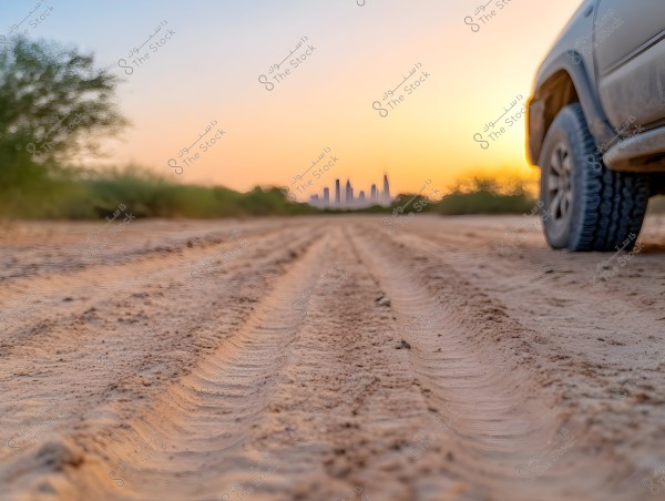 An image depicting part of a car on a sandy desert road at sunset. The car tire tracks lead towards the horizon where distant skyscrapers are visible among scattered desert plants. The sky is painted with vibrant shades of orange and yellow.