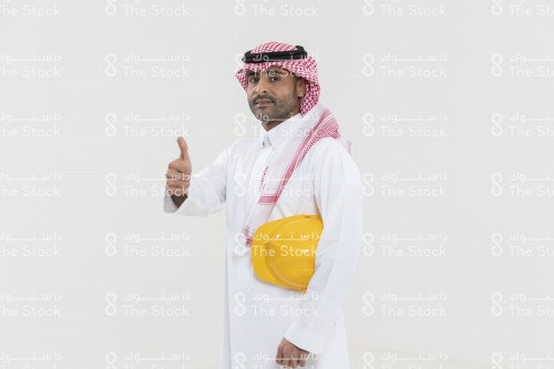 بورتريه صورة مقربة لمهندس سعودي يرتدي الثوب ويحمل تحت ذراعه خوذة المهندسين ويشير بيده علامة الإعجاب.