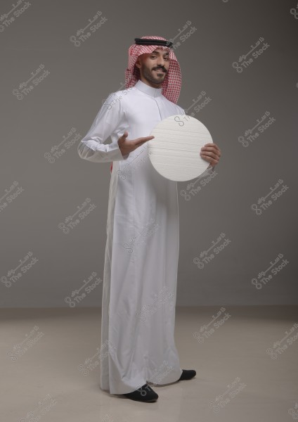 An image of a man standing against a gray background, wearing a white thobe and a red and white ghutra with a black agal, indicative of the Arabian Peninsula, possibly Saudi Arabia. He is holding a white circular board in his right hand and pointing at it with his left hand.