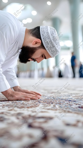 A man wearing a white thobe and a patterned cap is performing prayer inside a mosque. The focus is on the details of the carpet with traditional Arabic motifs. The mosque’s pillars are visible in the background, and the carpet extends across the floor.
