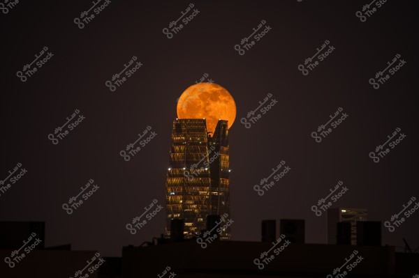 An image showing a bright orange full moon rising above the top of a modern building with several glass towers at night. The building is illuminated with lights and extends into the dark sky. In the foreground, faint silhouettes of other building rooftops are visible.