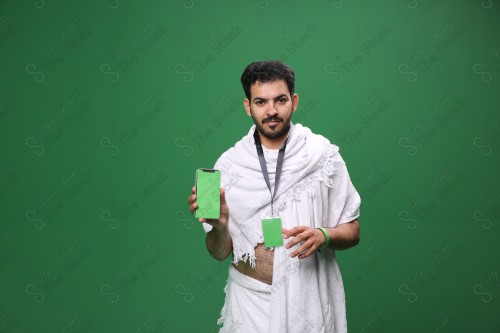 Portrait of a Saudi pilgrim on a green background wearing the Ihram to perform Hajj, carrying a mobile phone and reviewing the green health status