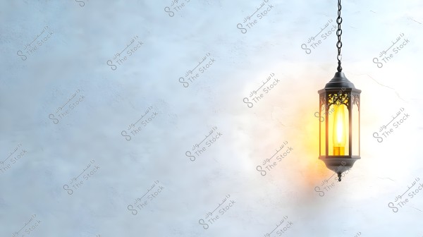 A hanging metal lantern with traditional Arabic decorative design, glowing with a warm yellow light against a smooth white wall background. The light emanating from the lantern casts a beautiful glow on the surrounding wall.