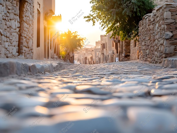 Image of a cobblestone street in a traditional village, flanked by old stone buildings. The sun is setting on the horizon, casting a golden glow that enhances the serene, antique atmosphere of the place. Some trees are visible on the sides of the street.