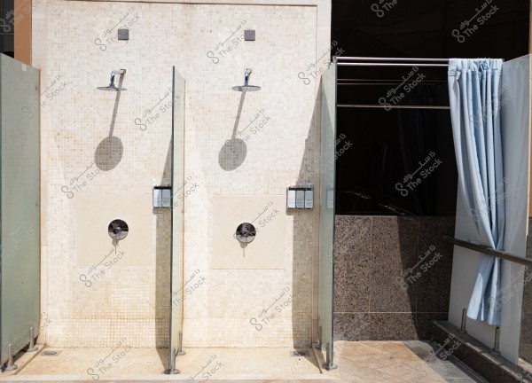 Two adjacent outdoor swimming showers with light beige tiles. Each shower features a metallic shower head and soap dispensers mounted on the walls. Transparent glass partitions separate the showers. Blue curtains are positioned next to them.