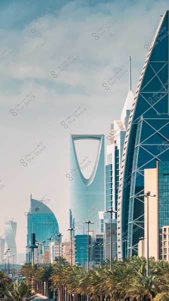 صورة لمجموعة من الأبراج الحديثة في مدينة الرياض, المملكة العربية السعودية. يظهر في الصورة برج المملكة بواجهته الزجاجية المميزة في الوسط, محاطاً بعدة ناطحات سحاب أخرى بتصميمات معمارية حديثة. تُظهر الصورة صفاً من أشجار النخيل على جانبي الطريق المؤدي إلى هذه الأبراج, في يوم صافٍ مع سماء زرقاء.