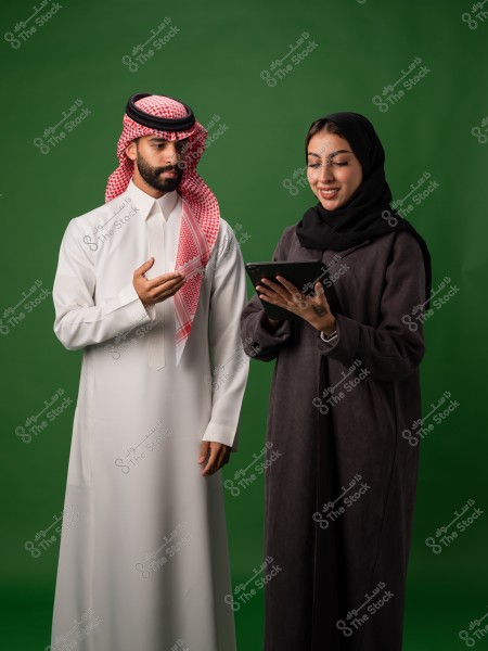 رجل وامرأة سعوديان يرتديان الملابس التقليدية يقفان أمام خلفية خضراء ويتفاعلان مع جهاز لوحي.