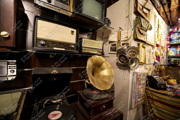 Image of a collection of old electronics in a shop or exhibition, including vintage radios of various sizes and a gold-colored gramophone. In the background, there are several hanging tools and bags, with multiple decorative details from a past era.