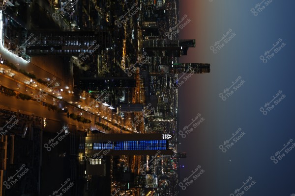 A night view of a cityscape illuminated by streetlights and glittering buildings. The image features a tall building with blue lights and a lighting pattern in the center, alongside a wide avenue filled with moving cars. The skyline shows a gradient of sky colors from dark to light as it fades into the night.