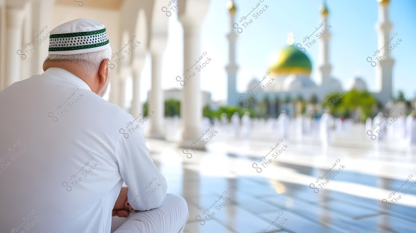 Image of a man sitting in traditional white clothing in the foreground, wearing a white cap with green stripes. In the background, there\'s a large mosque with a golden dome and white minarets, with several people in white clothing. The scene appears to be captured on a sunny day.