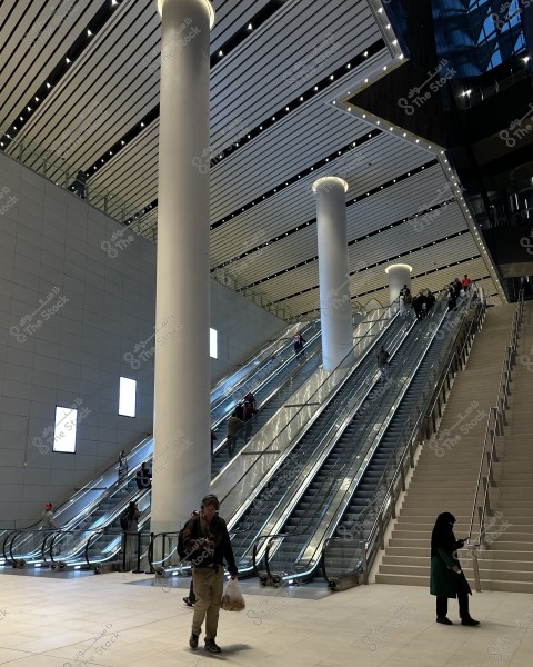 Image of a modern interior space featuring several large escalators and massive columns. Several people are using the escalators, most wearing winter clothing indicating cold weather. The overall design is modern and suggests a location like a shopping mall or large transit station. A woman in a dark long coat is in the foreground, and other individuals are dressed in casual attire.