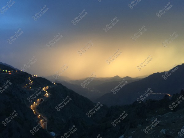 An image showing a winding road illuminated with yellow lights stretching through a range of mountains at sunset. The sky is a mix of blue and orange hues, while the mountains cover the distant view with their dark outlines.