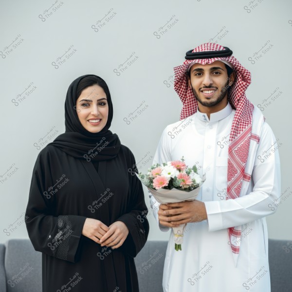 A photo of a man and a woman in traditional clothing. The woman is wearing a black abaya and headscarf, smiling. The man is wearing a white thobe and a red and white patterned shemagh, holding a bouquet of colorful flowers. The background is a grey wall.