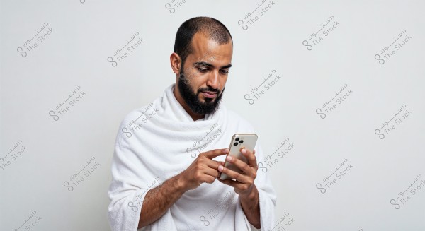 A portrait of a man wearing a white ihram, looking at and using his mobile phone with his hand. The background is plain white, and the image conveys a calm, contemporary scene.