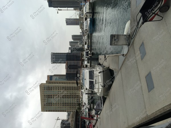 A view of a marina with multiple boats moored alongside a concrete dock. In the background, there are several high-rise buildings, some under construction, with cranes visible. The sky is overcast with clouds above.