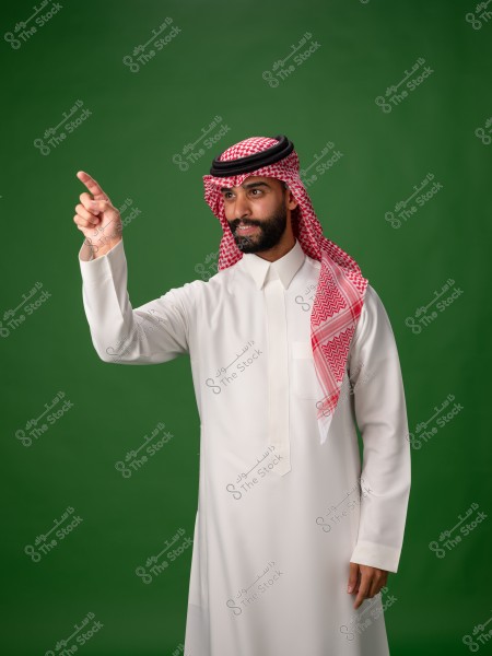 Portrait of a man wearing traditional Saudi attire, consisting of a white thobe and a red checkered headscarf (ghutra) with a black agal. He stands against a green background, pointing upwards with his finger.