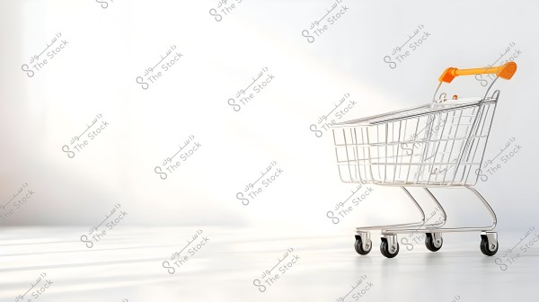 A metal shopping cart with orange handles placed on a white floor against a bright background. The cart appears empty and is positioned on the right side of the image, highlighting the shadows cast by the cart.
