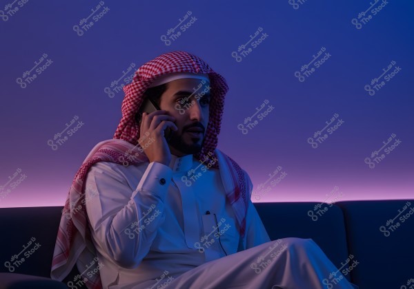 A portrait of a man wearing traditional Saudi attire, sitting on a couch while talking on the phone. He is dressed in a white thobe and a red and white checkered headscarf. The room has a purple backlight ambiance.