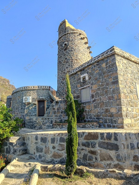 تظهر الصورة مبنى حجري قديم يتكون من برج دائري ومبنى مستطيل متصل, مع شجرة طويلة نحيفة في المقدمة. السماء زرقاء بالكامل والخلفية تظهر الطبيعة الجبلية. المبنى مبني بالكامل من الحجارة ويضم نافذة مكيفة وجدران متينة, مع تفاصيل معمارية مميزة.