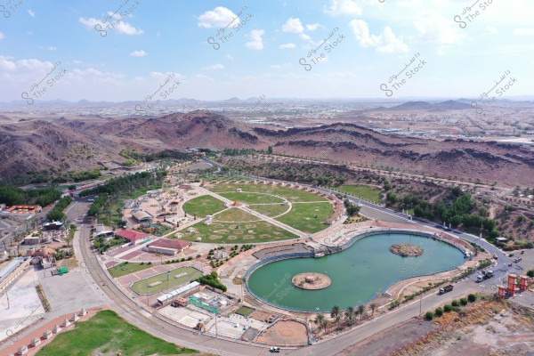 An aerial view of a large park situated in a mountainous area. The image shows expansive green spaces intersected by walking paths, along with a large circular lake featuring an island in the center. Paved roads and palm trees surround the park. In the background, a series of rocky mountains are visible, with the city skyline stretching into the distance under a blue sky with scattered clouds.