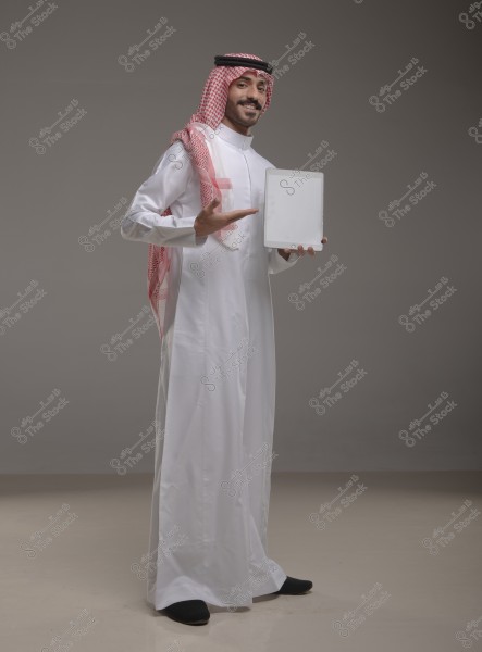 A portrait of a man standing in an empty studio wearing traditional Saudi attire, including a white thobe and a red and white checkered shemagh. The man holds a tablet in his right hand and points at it with his left hand while smiling at the camera.