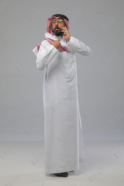 صورة لرجل يرتدي الثوب الأبيض التقليدي والشماغ المنقوش باللونين الأحمر والأبيض مع العقال الأسود, واقفاً وهو يتحدث عبر الهاتف المحمول. يبدو أنه في بيئة داخلية مع خلفية رمادية بسيطة.