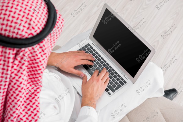 An image of a man wearing traditional Gulf attire, including a white thobe, red and white checkered headscarf, and black agal, working on a laptop. The laptop is open and the man appears to be typing. The background features a light-colored wooden floor.
