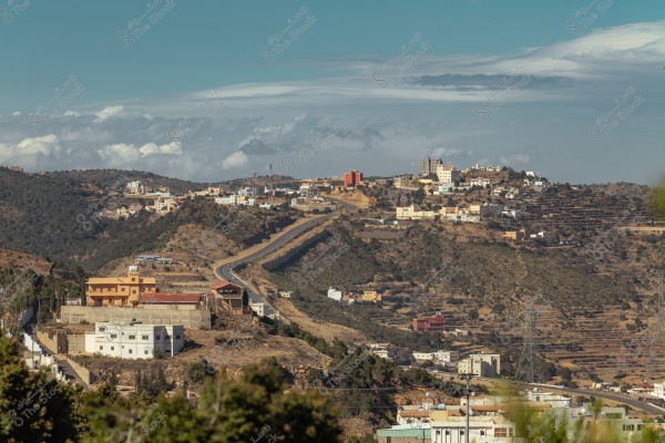 A scenic view showing a mountainous town with buildings scattered across the hills. A narrow winding road stretches between the hills. Green vegetation surrounds the area, and the sky is blue with some clouds on the horizon.