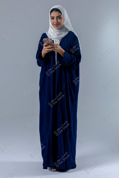 A portrait of a woman wearing a dark blue abaya and a white hijab. She is holding a mobile phone and smiling. The background is plain with a light gray color.