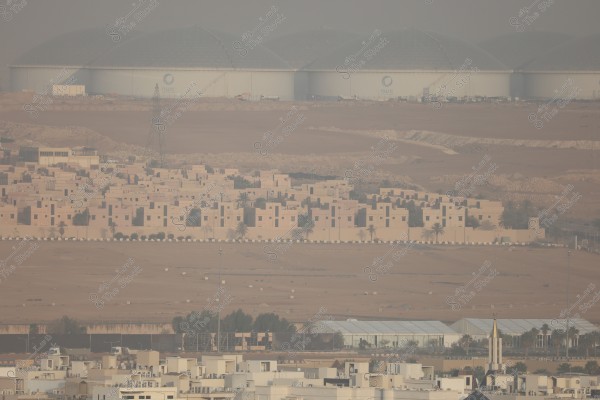 A view of a residential area in a desert environment. The foreground features rows of houses with traditional Arabic architecture surrounded by palm trees. In the background, there are large water tanks labeled with the National Water Company logo, with expansive desert plains and a slightly hazy skyline.