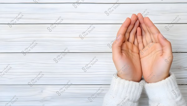 Two hands cupped together in a praying position on a white wooden surface. The sleeves are white and thick, resembling winter clothing.