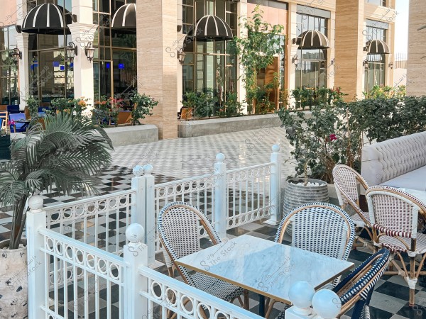 The image shows an outdoor café with elegant decor, featuring white and blue chairs arranged around a marble table. The exterior walls of the building are adorned with large glass windows topped with black and white striped awnings. Green plants and flowers add beauty to the setting, and there is a patterned floor with black and white tiles.