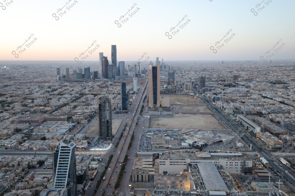 Aerial view of Riyadh city in Saudi Arabia, showcasing skyscrapers, modern buildings, and residential areas under a clear sky. The Kingdom Tower and other notable buildings are prominently visible, highlighting the city\'s urban expanse with roads and streets stretching between neighborhoods.