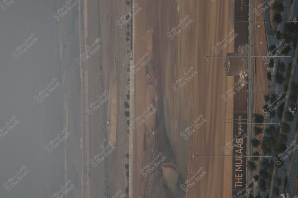 تُظهر الصورة منطقة شاسعة تحت الإنشاء, يبدو أنها موقع بناء مع بعض معدات الحفر الثقيلة والطريق المجاور. في الزاوية اليمنى يظهر جدار كبير عليه كتابة باللغة الإنجليزية تقول "THE MUKAAB A NEW ICON". المساحات المحيطة تبدو قاحلة مع وجود بعض الأشجار المتفرقة.