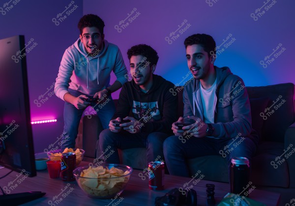Three young men sitting on a couch playing video games with controllers. The room is lit with blue and purple neon lighting. There are bowls of potato chips and cans of soft drinks on the table in front of them. The men are dressed in casual clothing, such as jeans and hoodies.