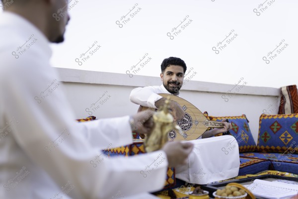 شاب يجلس في مجلس عربي تقليدي يمسك بآلة العود وعليه ابتسامة, بينما يقدم له شخص آخر القهوة العربية.