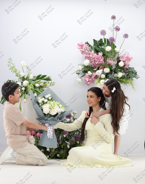 The image shows a woman in a light-colored long dress sitting and smiling while holding a bouquet of white and green flowers. A child kneels in front of her presenting the flowers, and a small girl stands behind her, smiling. The background is filled with floral arrangements of pink, purple, and white flowers.