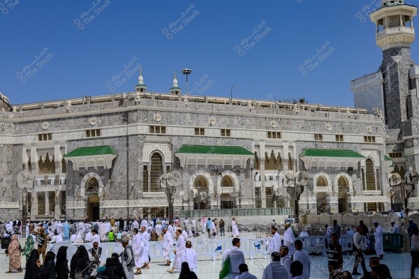 صورة تظهر ساحة المسجد الحرام في مكة المكرمة, المملكة العربية السعودية, حيث يتجمع عدد كبير من الناس بملابس إحرام بيضاء, ويظهر جزء من الكعبة المشرفة في الخلفية, مع تفاصيل معمارية إسلامية على الجدران. السماء زرقاء وصافية في الأعلى.